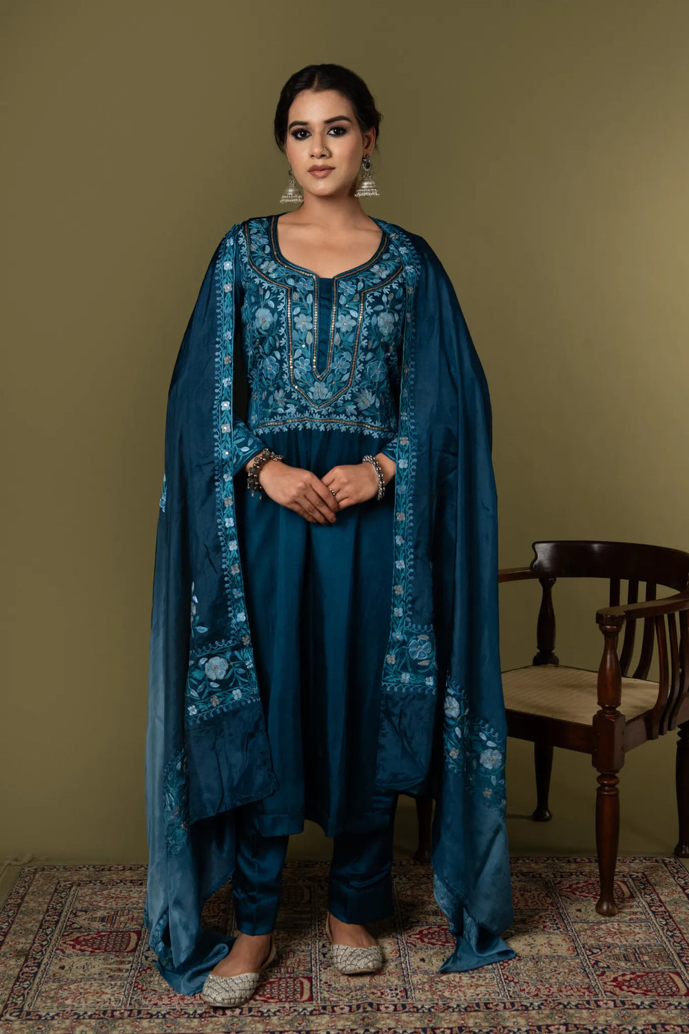 #TealBlue Woman wearing a blue embroidered traditional outfit standing against a plain background Woman in a blue traditional outfit with a cape standing on a patterned rug against a green wall.