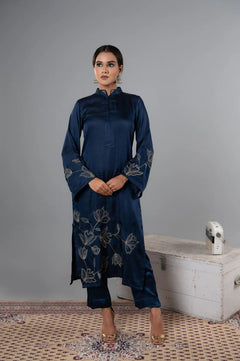Woman wearing a dark blue embroidered outfit standing against a gray wall.